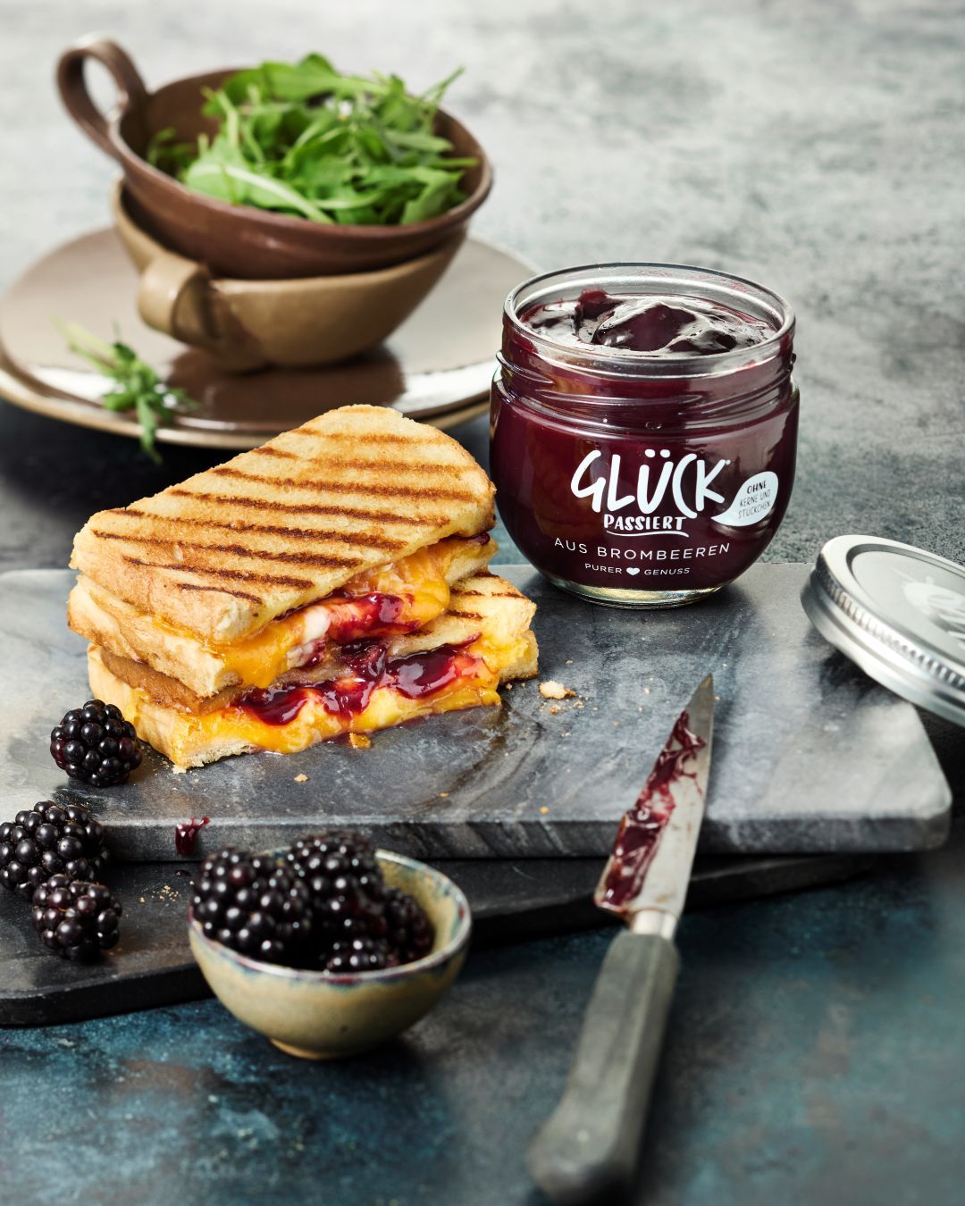 Grilled Cheese Sandwich mit Käse und GLÜCK passiert aus Brombeeren. Daneben ein offenes Glas GLÜCK passiert aus Brombeeren, einige Brombeeren und ein Messer.