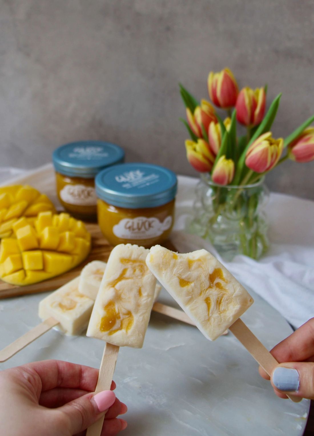 Mango Joghurt Eis am Stiel, gehalten von zwei Händen. Im Hintergrund zwei Gläser GLÜCK passiert mit Leichtigkeit aus Mango.