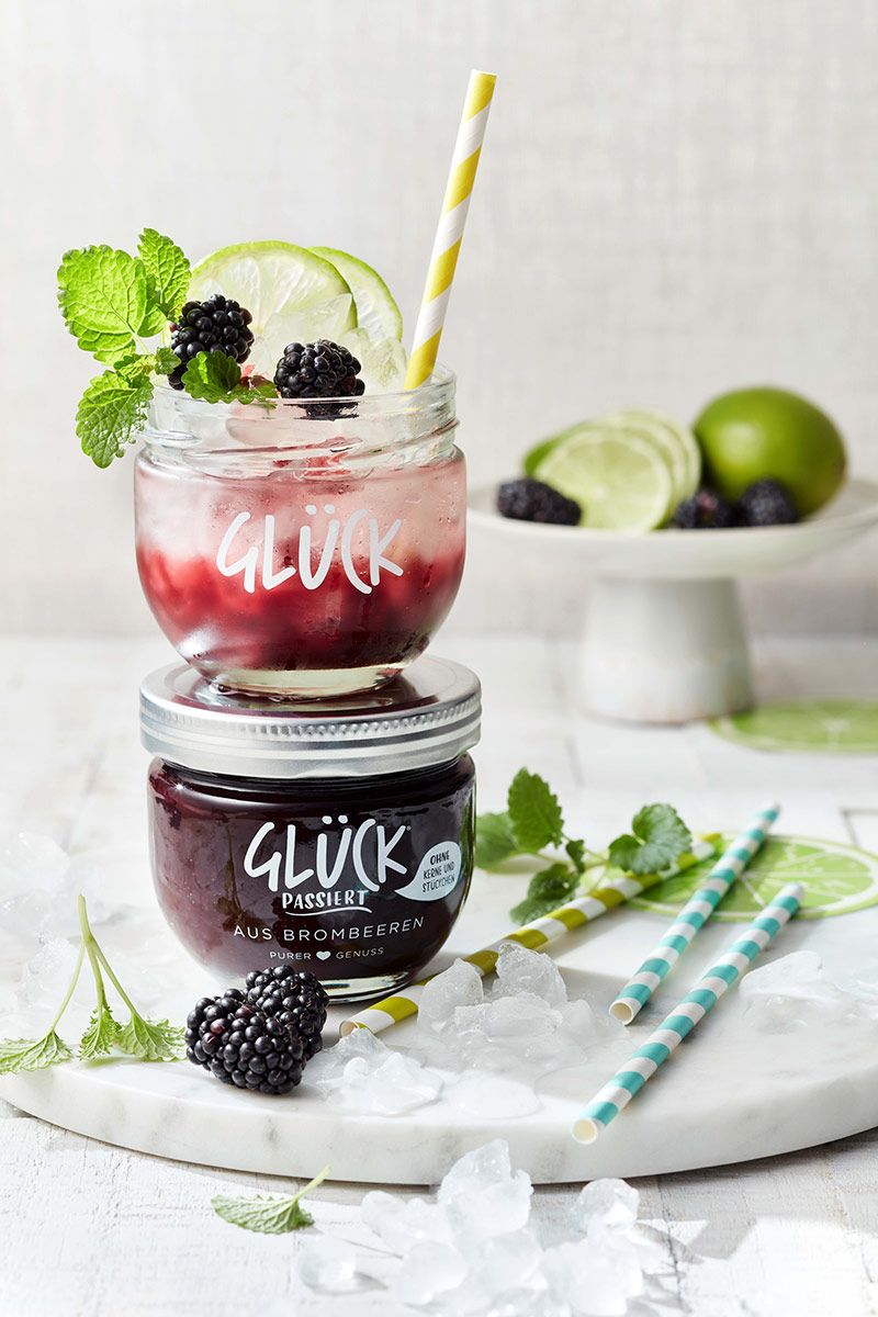 Brombeer Bramble, gefüllt in ein leeres GLÜCK passiert aus Brombeeren. Der Drink ist garniert mit frischen Beeren und Zitronenmelisse und steht auf einem vollen Glas GLÜCK passiert aus Brombeeren. Daneben liegen Eis, Zitronenmelisse und Strohhalme.