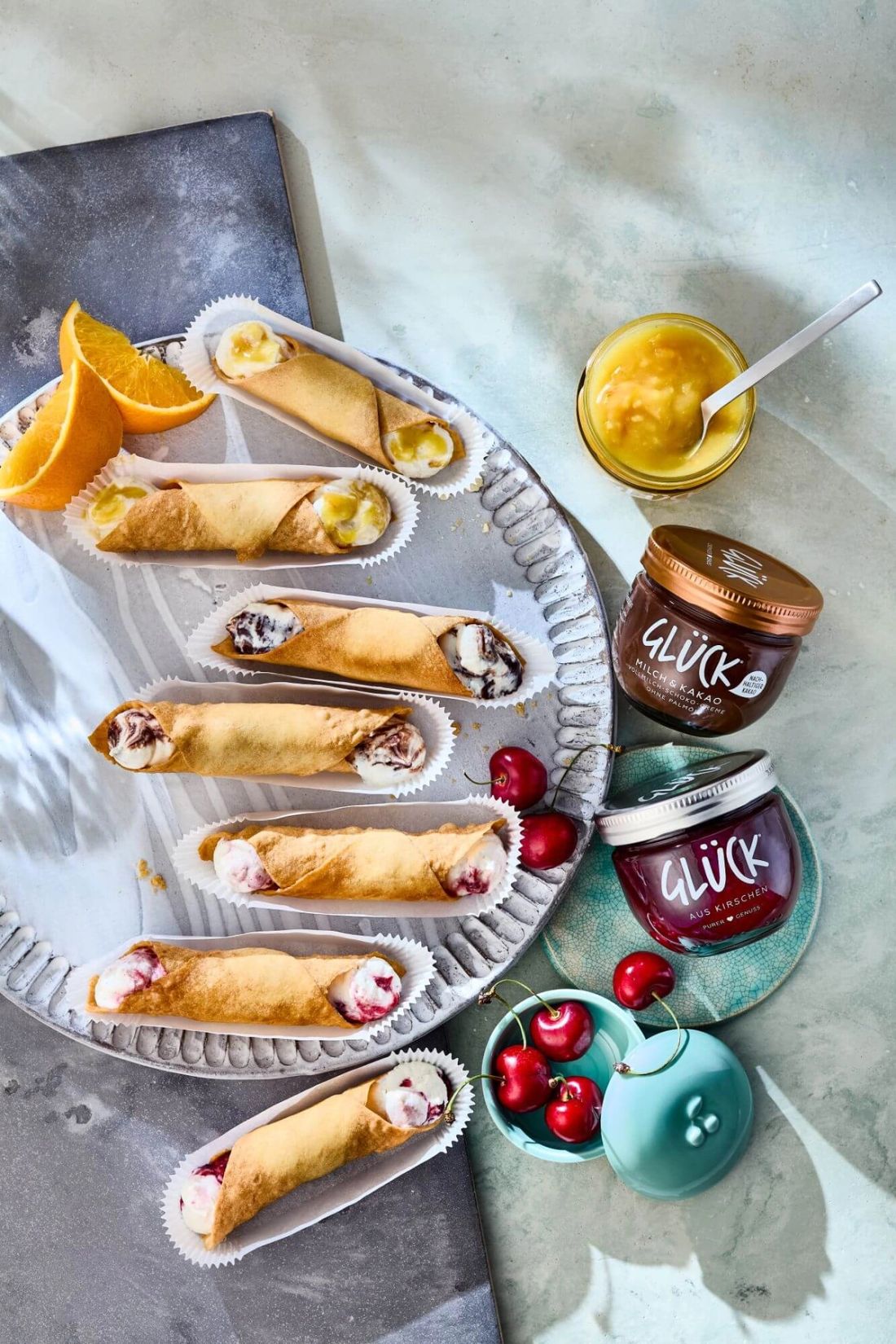 Cannoli mit Ricotta-Fruchtfüllung neben Kirschen und Orangen sowie einem Glas GLÜCK Milch & Kakao und GLÜCK aus Kirschen.