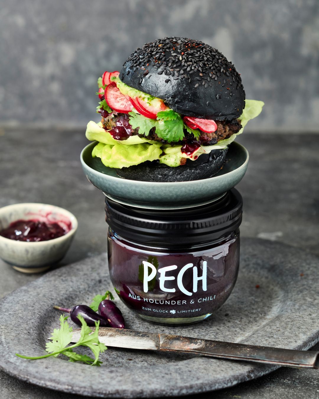 Burger mit schwarzen Brötchen, Black-Bean-Patties und Holunder-Chili-Chutney. Der Burger steht auf einem Glas PECH aus Holunder & Chili. Im Hintergrund mehr Chutney.