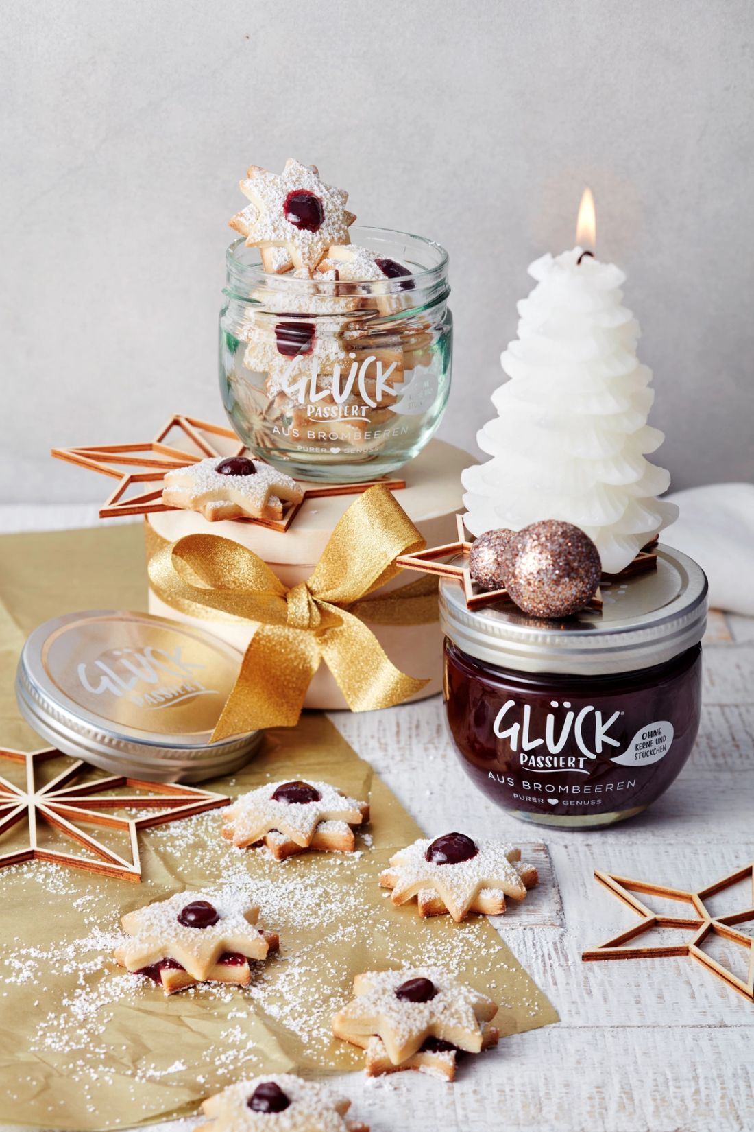 Linzersterne mit GLÜCK aus Brombeeren, darum Weihnachtsdekoration mit einer Schleife und einer Tannenbaumkerze. Daneben steht ein Glas GLÜCK passiert aus Brombeeren.