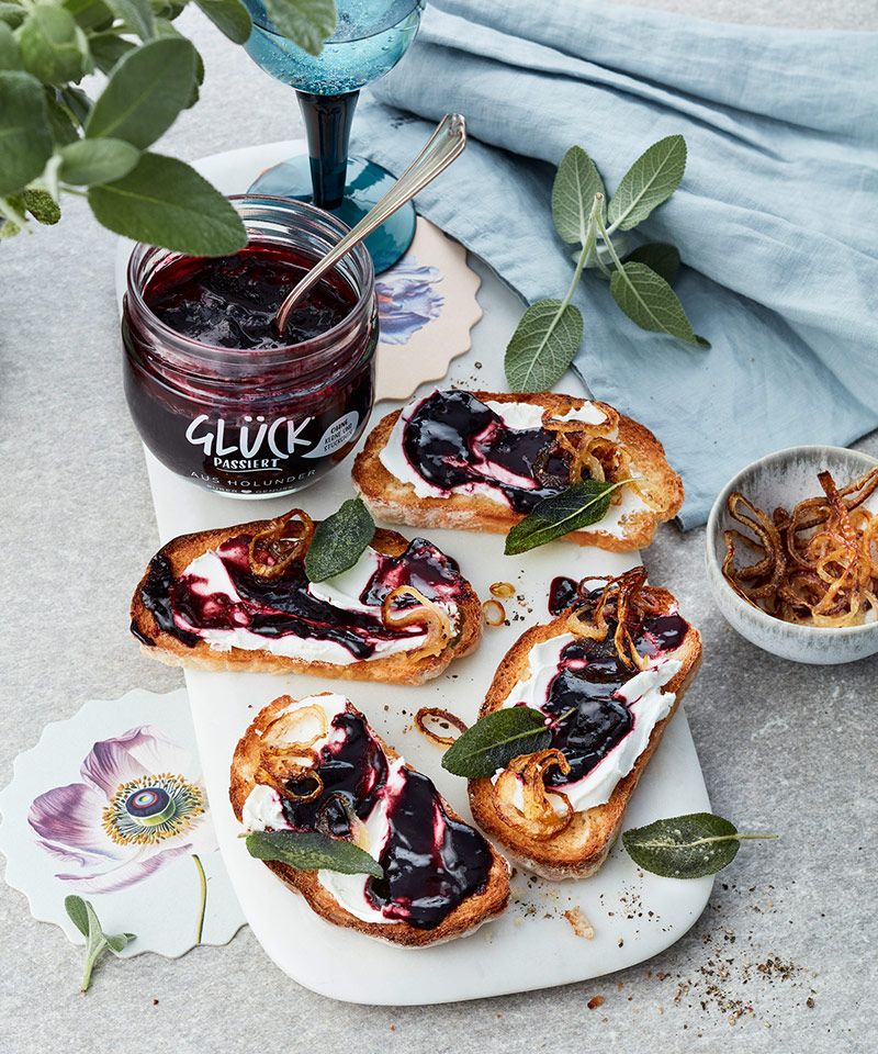 Bruschetta mit Ziegenfrischkäse und GLÜCK passiert aus Holunder auf einem Brett. Garniert mit Salbeiblättern. Daneben steht ein volles Glas GLÜCK passiert aus Holunder und eine Schüssel mit karamellisierten Zwiebeln.
