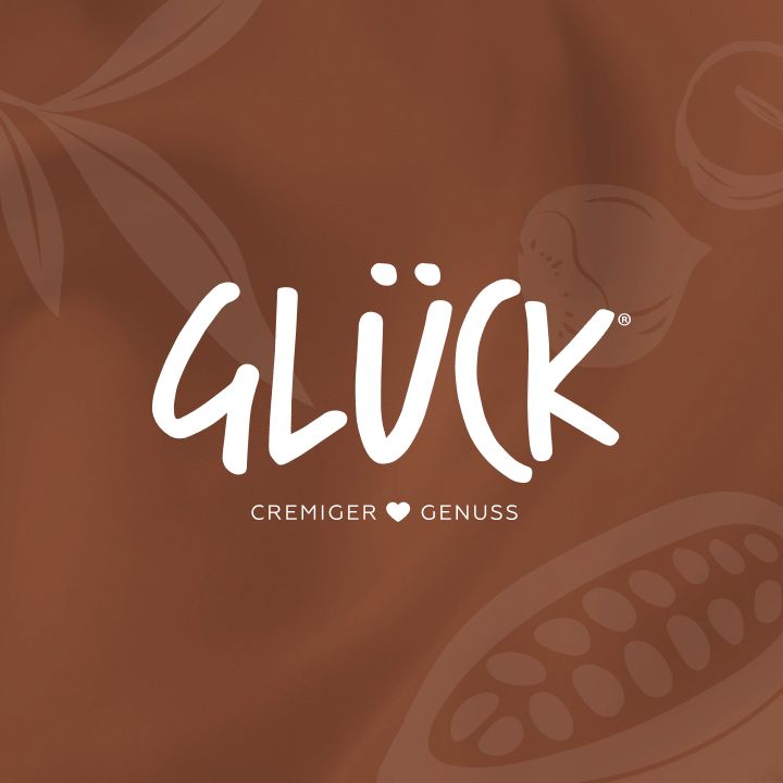 Brauner Hintergrund mit hellbraunen Grafiken von Kakaobohnen und Nüssen. In der Mitte in weiß das Logo "GLÜCK cremiger Genuss" mit einem kleinen Herz zwischen den Worten "cremig" und "Genuss".