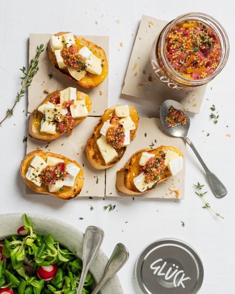Orangen-Chutney auf Baguette mit Camembert. Neben den Baguettescheiben liegt ein Löffel, daneben steht ein volles Glas Chutney aus GLÜCK aus Orangen. Daneben als Beilage Feldsalat mit Radieschen.