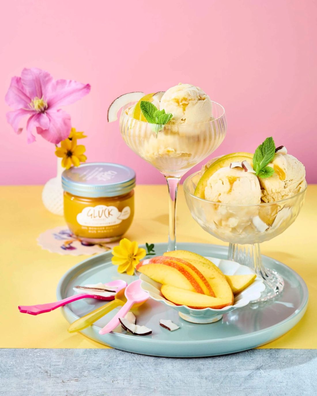 Mango-Kokos Nicecream im Glas. Daneben Mangostücke und ein Glas GLÜCK passiert mit Leichtigkeit aus Mango.