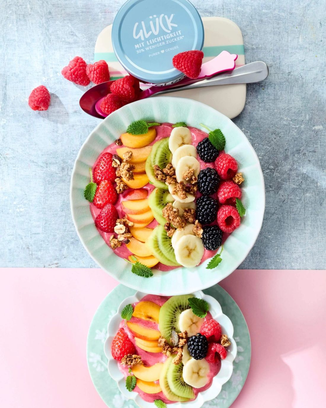 Himbeeren Smoothie Bowl in einer Schüssel mit Früchten. Daneben ein Glas GLÜCK mit Leichtigkeit aus Himbeeren.