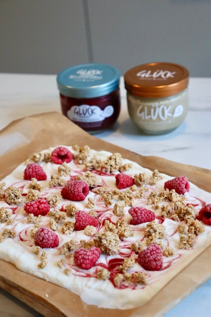 Fruchtiger Joghurt Bark mit Himbeeren und Granola. Dahinter zwei Gläser GLÜCK.