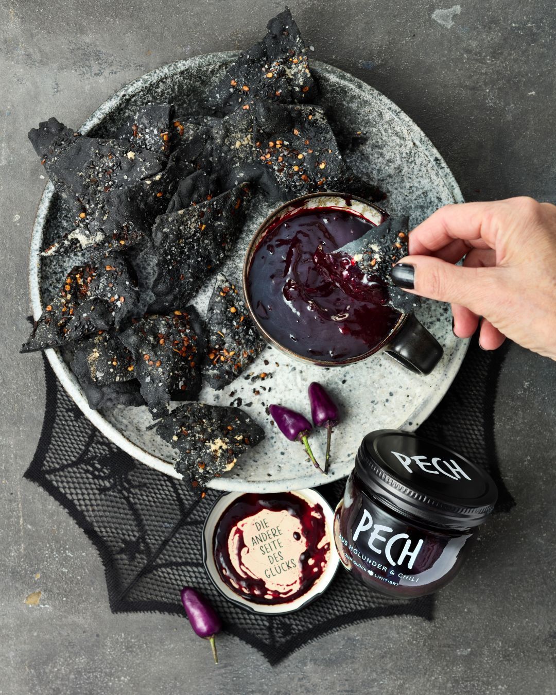Schwarze Tortilla-Chips mit Holunder-Chili-Dip. Eine Hand dippt einen Chip in den Dip. Daneben ein Glas PECH aus Holunder & Chili. Darum verteilt Halloween-Deko.