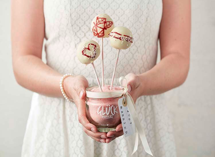 Eine Person im weißen Kleid hält ein GLÜCK Glas, in dem rosafarbener Zucker ist. In das Glas gesteckt sind weiße Cake Pops mit roter Verzierung. An dem Glas hängt ein Band mit einem Schild mit der Aufschrift "JUST MARRIED"