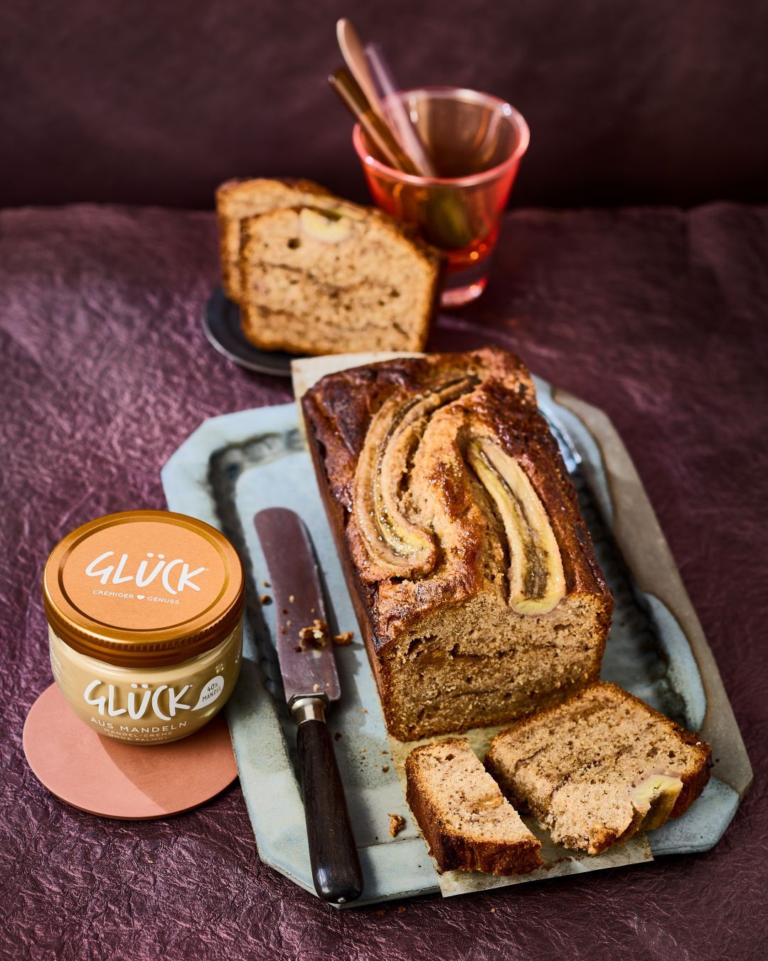 Bananenbrot auf einem Brett mit Messer und einem Glas GLÜCK aus Mandeln daneben.