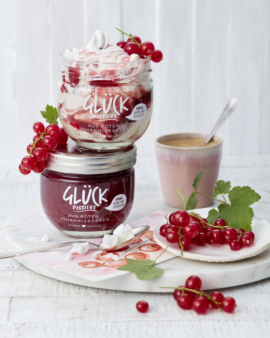Johannisbeere Eton Mess im GLÜCK Glas, garniert mit Johannisbeeren. Daneben ein Glas GLÜCK passiert aus roten Johannisbeeren.