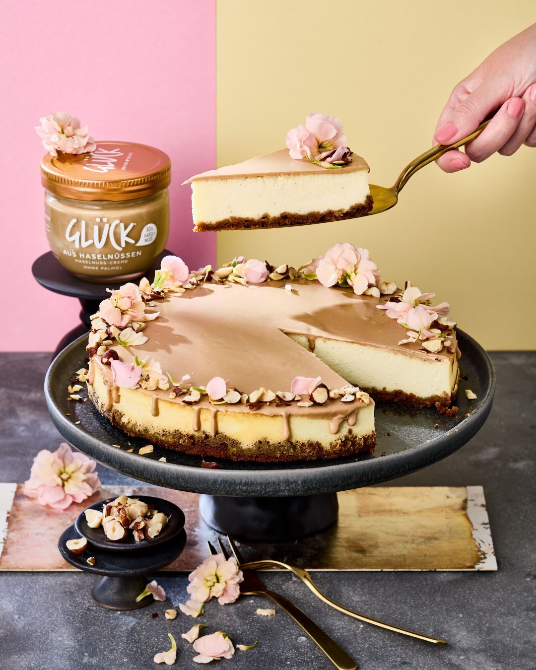Käsekuchen mit Haselnusscremespiegel und Blumen zur Deko. Ein Stück Kuchen wird angehoben.