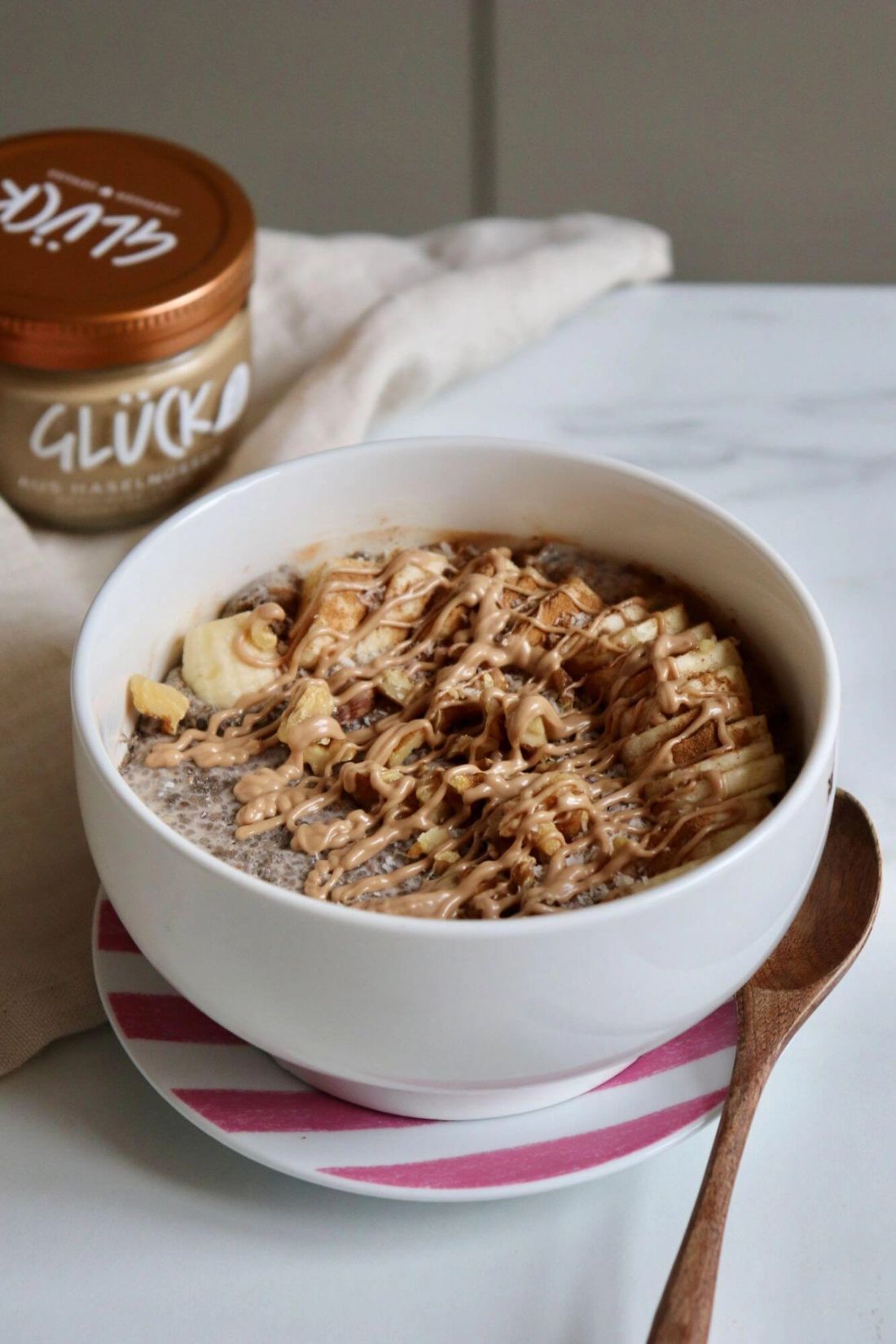 Bananen Chia Pudding in einer Schüssel mit Löffel. Daneben ein Glas GLÜCK aus Haselnüssen.