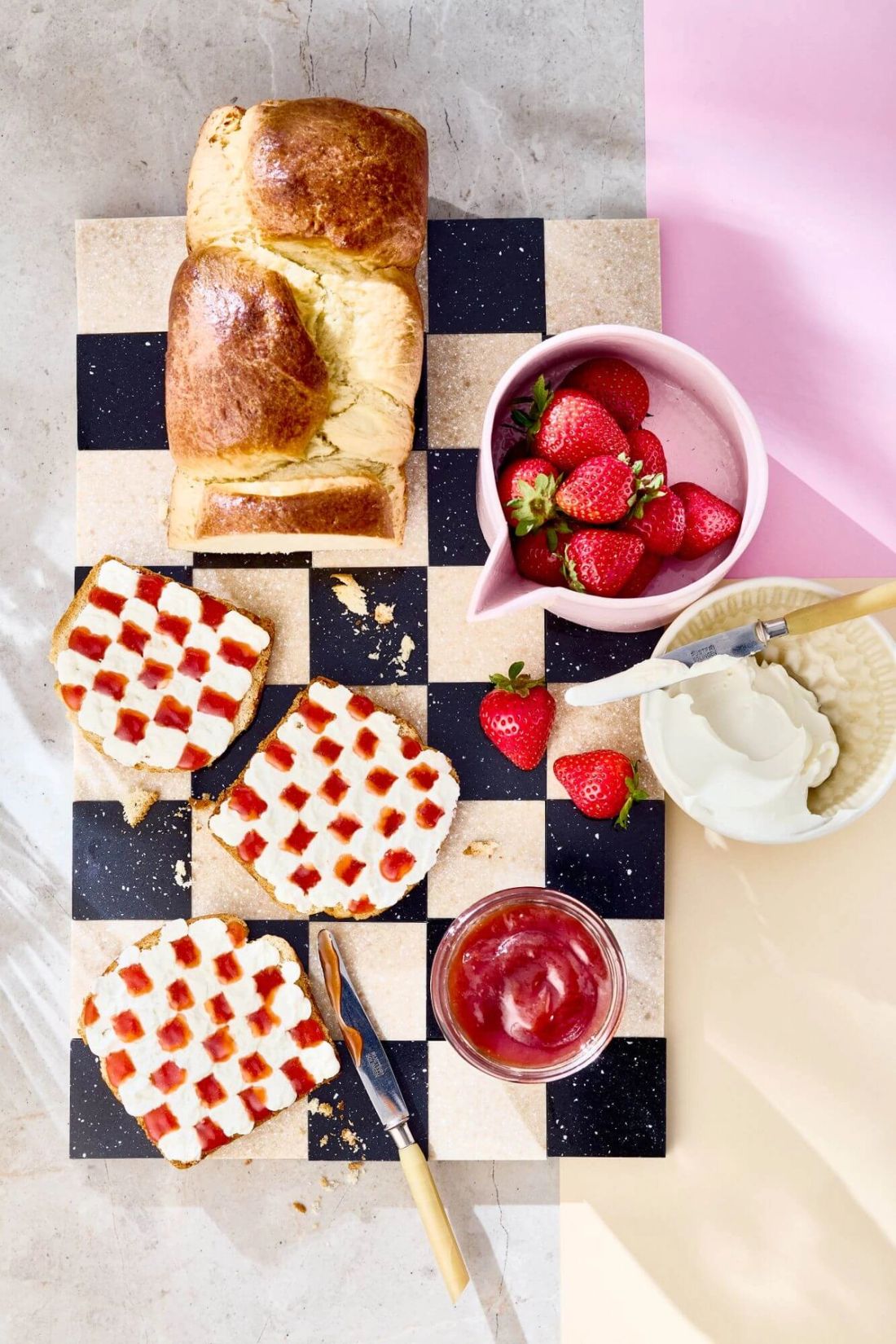 Schneidebrett mit Brioche, Toast und Erdbeeren. Frischkäse und Marmelade auf Toast sind im Schachbrettmuster angerichtet.