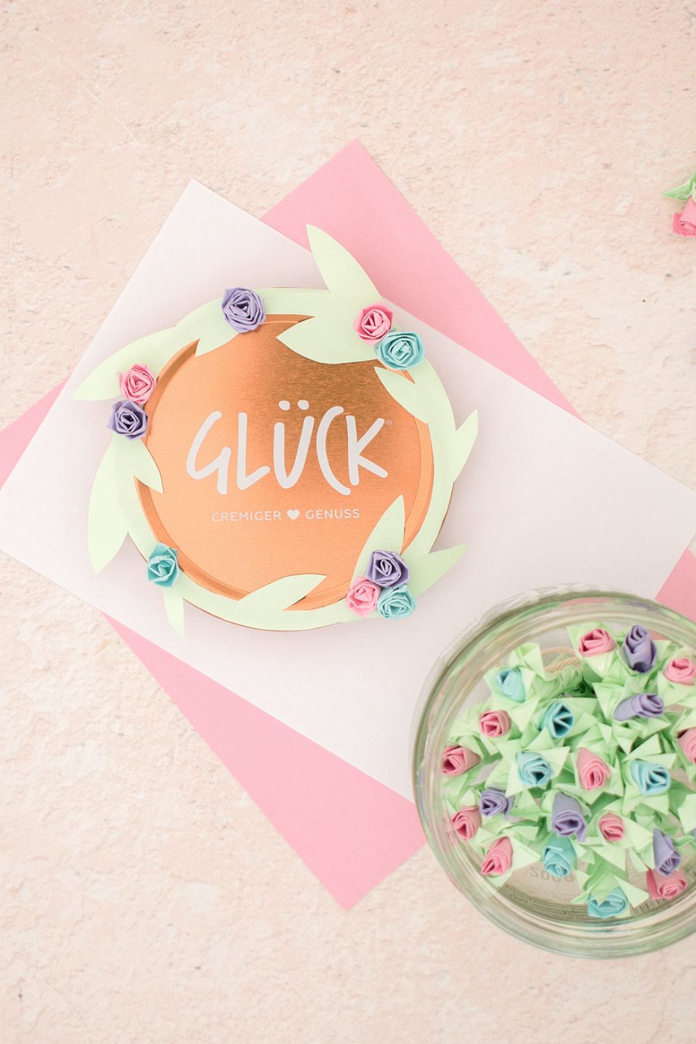 Eine Geschenkidee zum Muttertag. In einem leeren GLÜCK Glas liegen viele bunte Papierblumen mit rosa, lila und blauen Blüten. Der Deckel das Glases wurde mit einem grünen Rand in Form von Blättern verziert, an den ebenfalls bunte Blüten geklebt wurden.