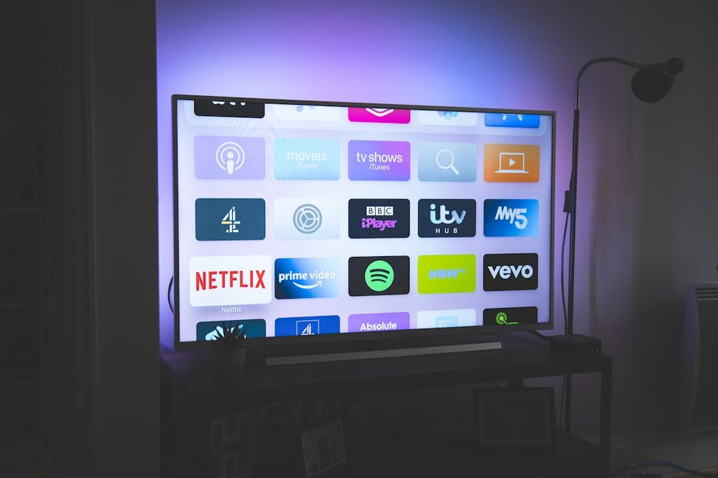 The image features a modern television displaying various streaming service icons on its screen. The setting has ambient lighting that enhances the visual appeal of the display. — Credit: Photo by Nicolas J Leclercq on Unsplash