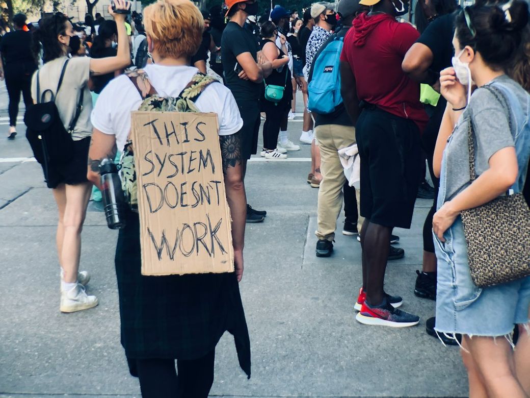 A demonstrator stands amidst a crowd of protesters, wearing a sign on their back that reads 'THIS SYSTEM DOESN'T WORK.' The setting suggests a social movement or protest related to systemic issues, with various individuals present and holding signs. — Credit: Photo by Tarun Girish on Unsplash