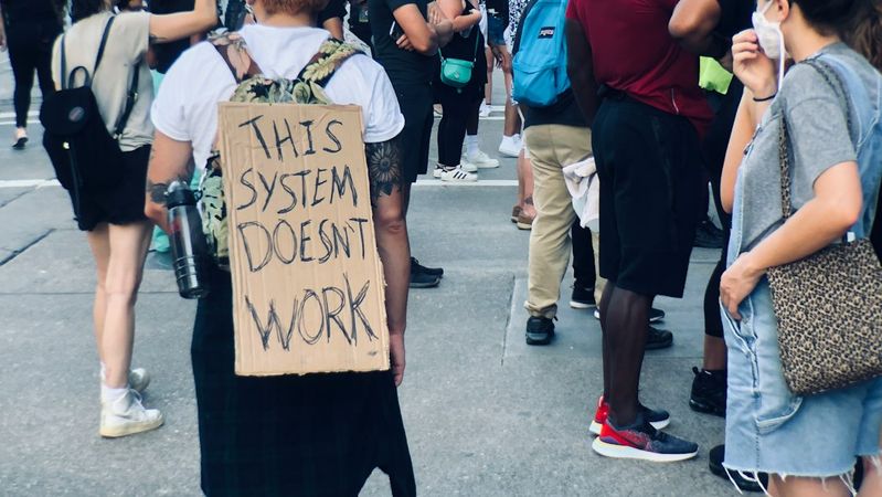 A demonstrator stands amidst a crowd of protesters, wearing a sign on their back that reads 'THIS SYSTEM DOESN'T WORK.' The setting suggests a social movement or protest related to systemic issues, with various individuals present and holding signs.