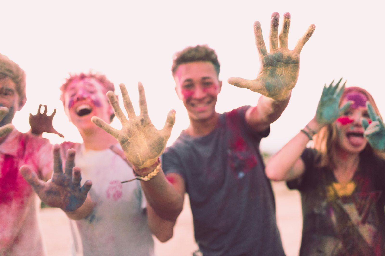A group of four young individuals engaging in a playful moment outdoors, covered in colorful paint. They are smiling and showing their painted hands towards the camera, conveying a sense of joy and creativity.
