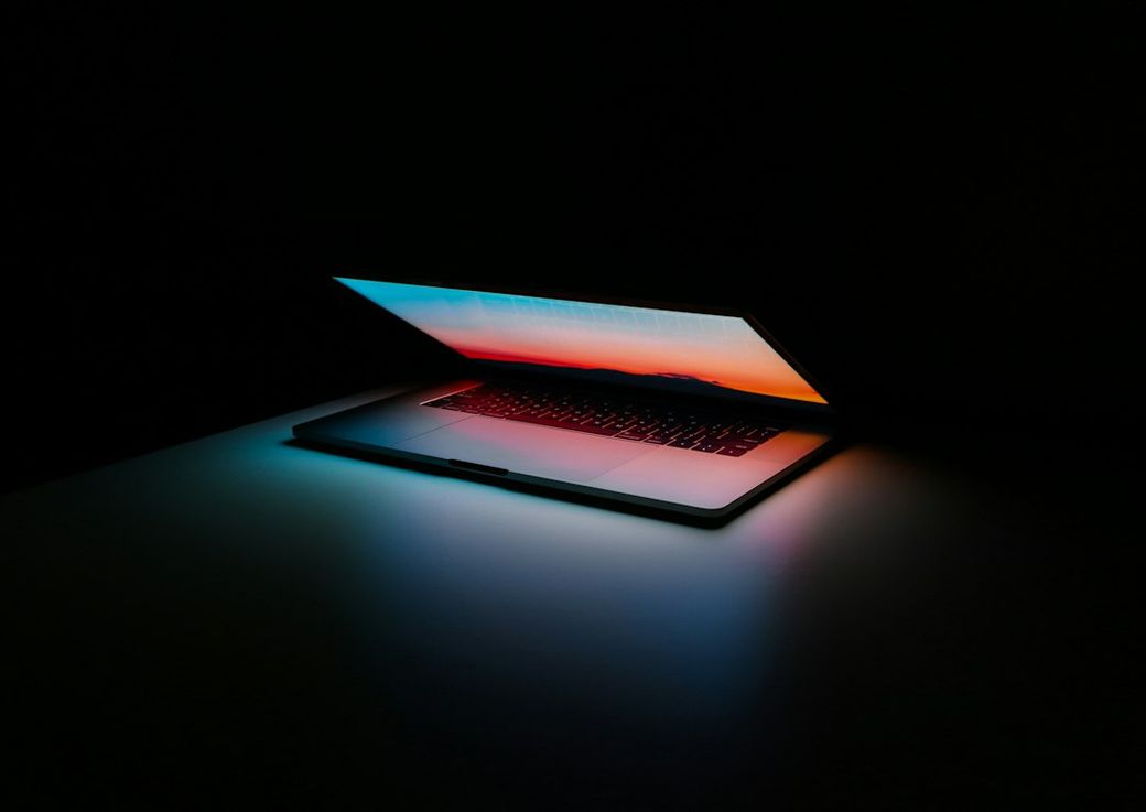 A laptop with a brightly lit screen displaying a colorful sunset against a dark background. The device is positioned on a surface, emphasizing its sleek design. — Credit: Photo by Ales Nesetril on Unsplash