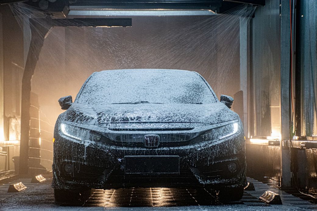 A car wash scene featuring a Honda vehicle being cleaned with soap and water. The image captures the car partially obscured by a foam spray during the washing process. — Credit: Photo by Zulfahmi Khani on Unsplash