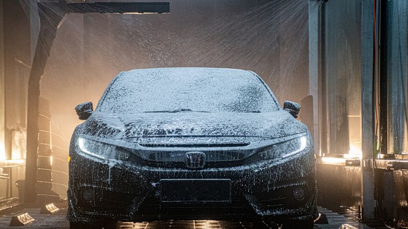 A car wash scene featuring a Honda vehicle being cleaned with soap and water. The image captures the car partially obscured by a foam spray during the washing process.