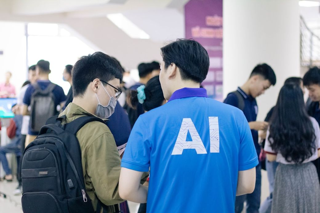 The image captures a busy indoor setting where a group of people are engaged in conversation and activities related to technology and artificial intelligence. Two individuals are prominently featured in the foreground, with one wearing a blue shirt that has 'AI' printed on the back. — Credit: Photo by Nguyen Dang Hoang Nhu on Unsplash