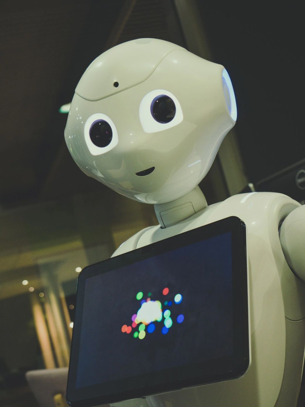 The image features a humanoid robot with a friendly expression, designed for interaction. The robot is positioned in a setting that suggests a technology-focused environment, likely for demonstration or user engagement purposes. — Credit: Photo by Owen Beard / Unsplash