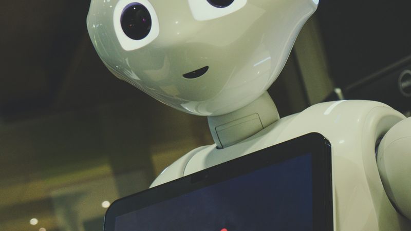 The image features a humanoid robot with a friendly expression, designed for interaction. The robot is positioned in a setting that suggests a technology-focused environment, likely for demonstration or user engagement purposes.