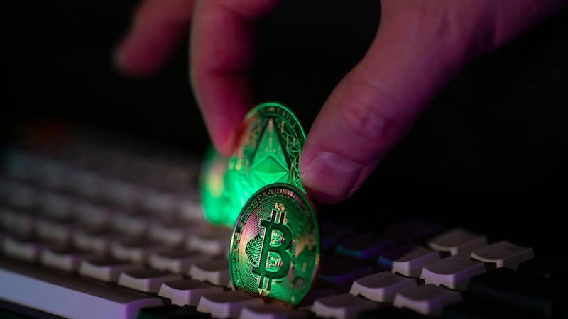 A close-up image showing a hand placing a glowing Bitcoin and Ethereum cryptocurrency coin on a keyboard. The illuminated coins reflect the digital nature of cryptocurrency and its interaction with technology.