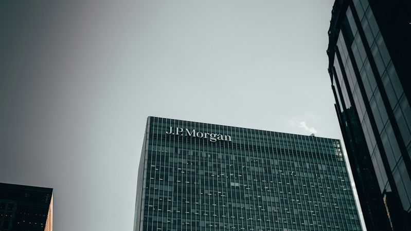 The image features the towering glass facade of a corporate building, prominently displaying the name 'J.P. Morgan' at the top. It is captured from a low angle, creating an imposing view against a clear sky.