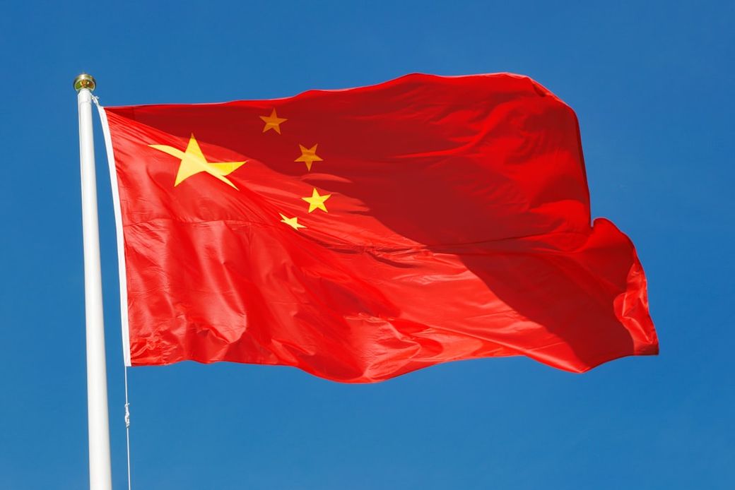 The image depicts the flag of China waving against a clear blue sky. The flag features a bright red background with five yellow stars, symbolizing the unity of the Chinese people. — Credit: Photo by CARLOS DE SOUZA on Unsplash