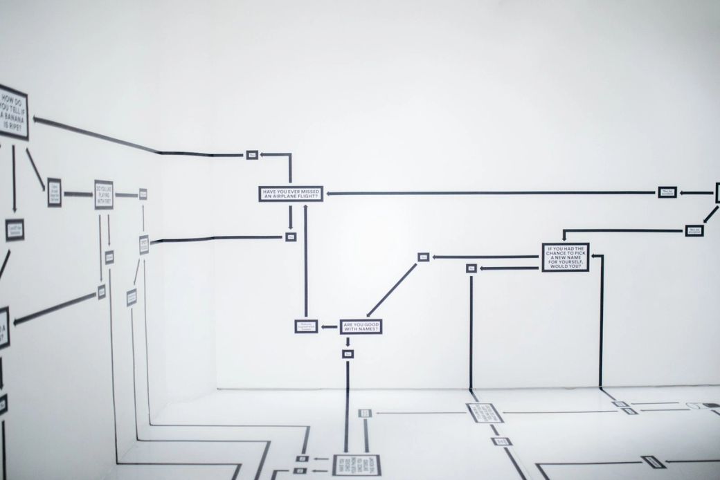The image depicts a minimalist white wall adorned with a series of black flowcharts and diagrams. The flowcharts appear to illustrate various processes or decision-making paths in a clear and structured manner. — Credit: Photo by Hanna Morris / Unsplash
