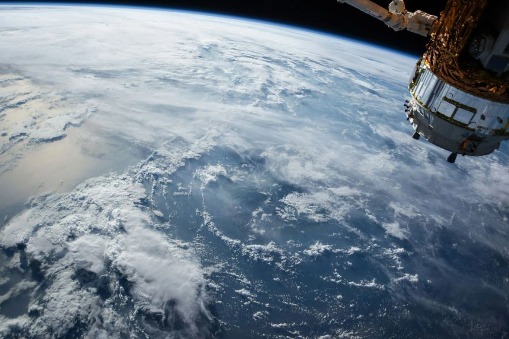 The image depicts a view of Earth from space, showcasing the curvature of the planet and its cloud cover. A section of a spacecraft can be seen in the foreground, highlighting human technology in orbit. — Credit: Photo by NASA / Unsplash