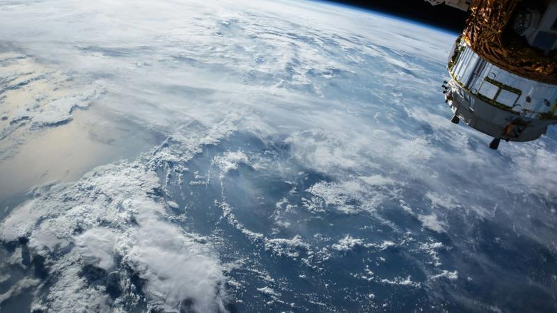 The image depicts a view of Earth from space, showcasing the curvature of the planet and its cloud cover. A section of a spacecraft can be seen in the foreground, highlighting human technology in orbit.