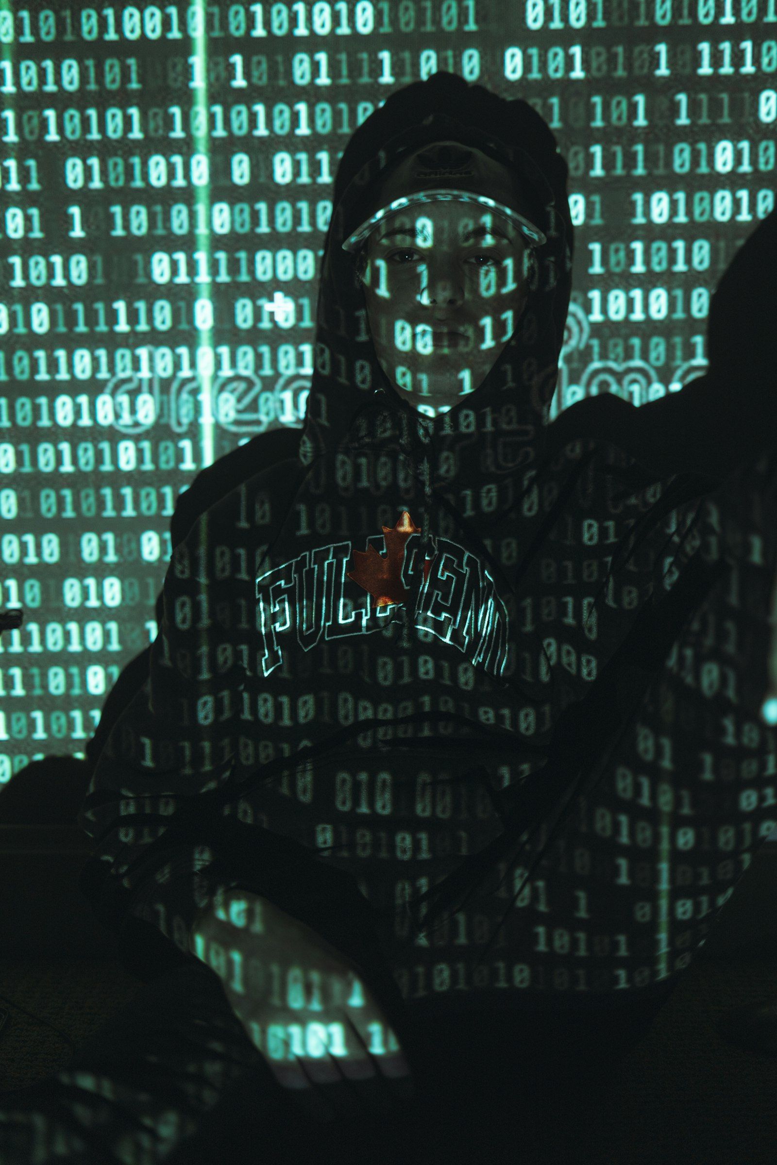 A person wearing a hoodie stands against a backdrop of green binary code, with their face illuminated by the projected numbers. The image evokes themes of technology and digital transformation.