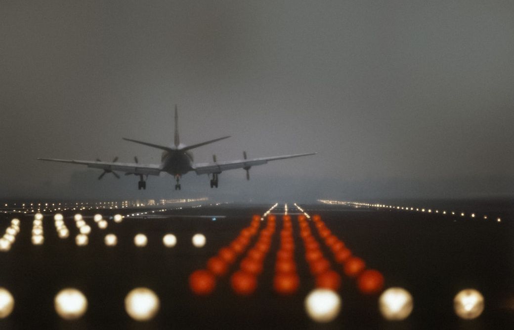 An airplane approaches a runway during twilight, illuminated by runway lights. The image captures a blurred view of the aircraft against a backdrop of low visibility conditions. — Credit: Photo by Amsterdam City Archives on Unsplash
