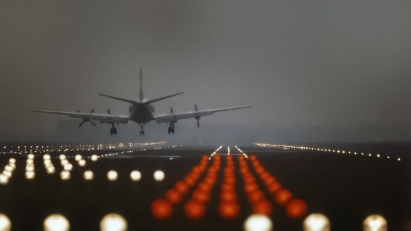 An airplane approaches a runway during twilight, illuminated by runway lights. The image captures a blurred view of the aircraft against a backdrop of low visibility conditions.