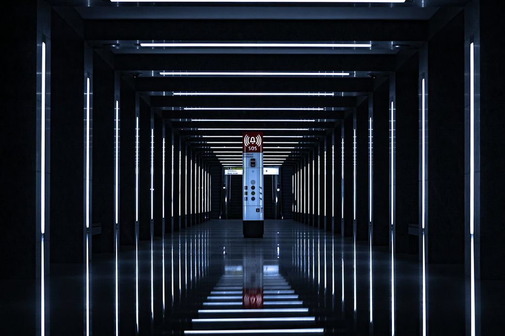 The image features a modern, illuminated corridor with parallel lines of light illuminating the path. At the end of the corridor is an elevator or lift, creating a stark visual focus against the dark surroundings. — Credit: Photo by Samuele Macauda on Unsplash