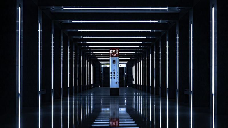 The image features a modern, illuminated corridor with parallel lines of light illuminating the path. At the end of the corridor is an elevator or lift, creating a stark visual focus against the dark surroundings.