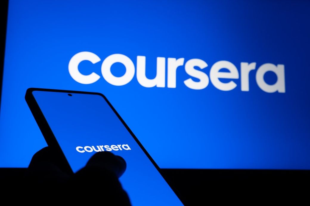 A person holds a smartphone in front of a screen displaying the Coursera logo. The background features a bright blue color, emphasizing the online learning platform's branding. — Credit: Photo by Zulfugar Karimov on Unsplash