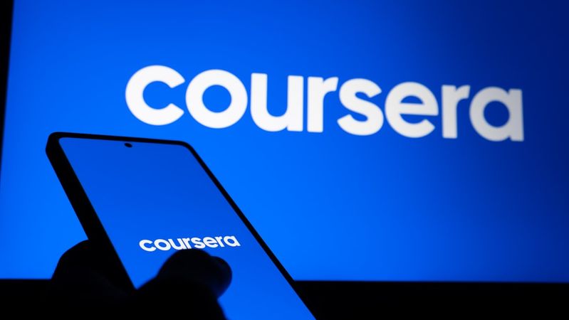 A person holds a smartphone in front of a screen displaying the Coursera logo. The background features a bright blue color, emphasizing the online learning platform's branding.