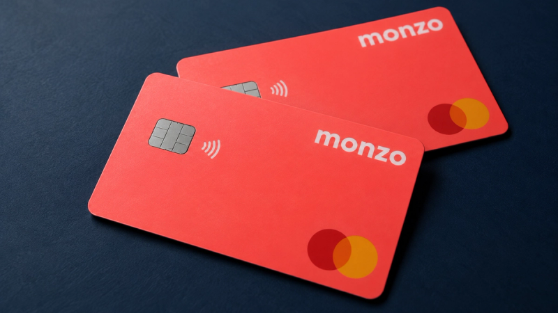The image features two Monzo debit cards prominently displayed on a dark surface. The cards are characterized by a bright red color, with the Monzo logo and Mastercard emblem visible, highlighting the brand's focus on modern banking solutions.