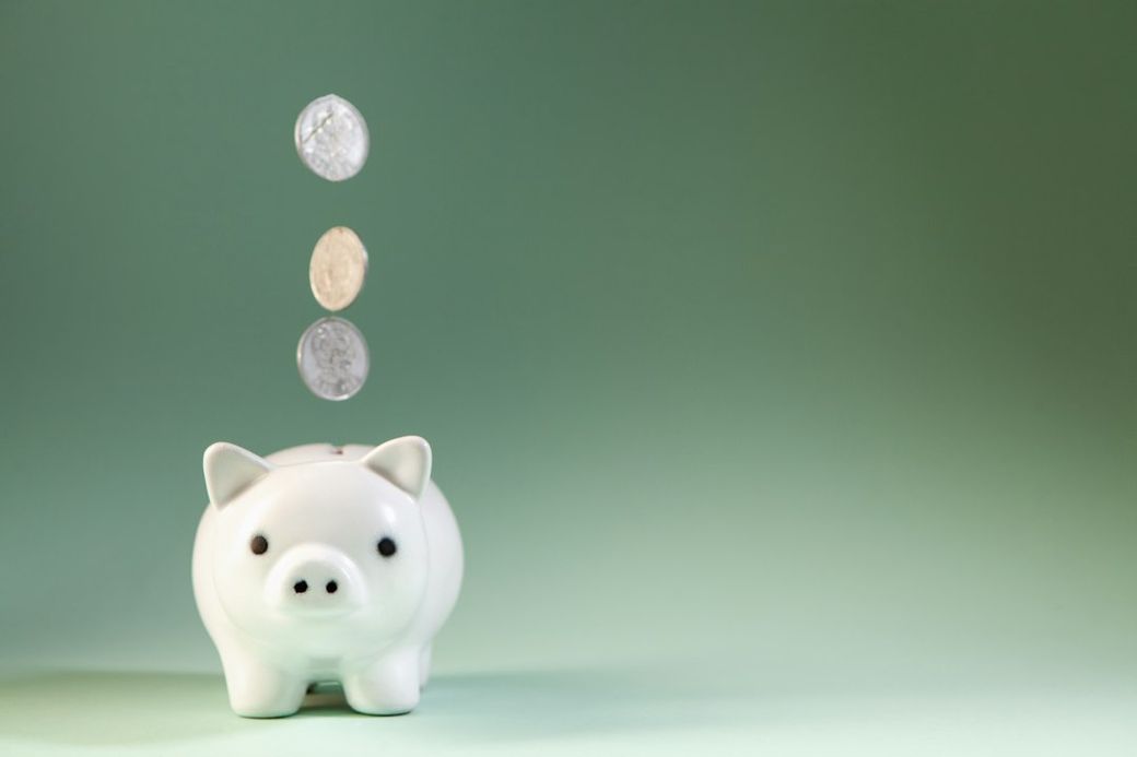 A white piggy bank is shown against a soft green background, with three coins appearing to drop into it from above. The image conveys themes of saving and finance. — Credit: Photo by Sasun Bughdaryan on Unsplash