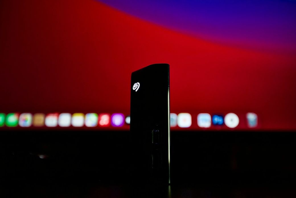 The image features a black external hard drive illuminated by a small light, positioned in the foreground against an out-of-focus backdrop of a bright red screen and various digital icons. This setup suggests a tech-centric environment, possibly related to digital storage or computing. — Credit: Photo by Ian Powell / Unsplash