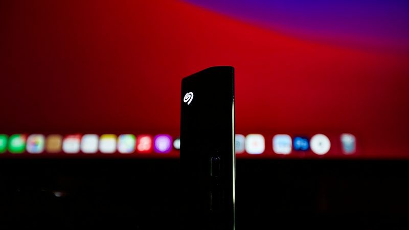 The image features a black external hard drive illuminated by a small light, positioned in the foreground against an out-of-focus backdrop of a bright red screen and various digital icons. This setup suggests a tech-centric environment, possibly related to digital storage or computing.