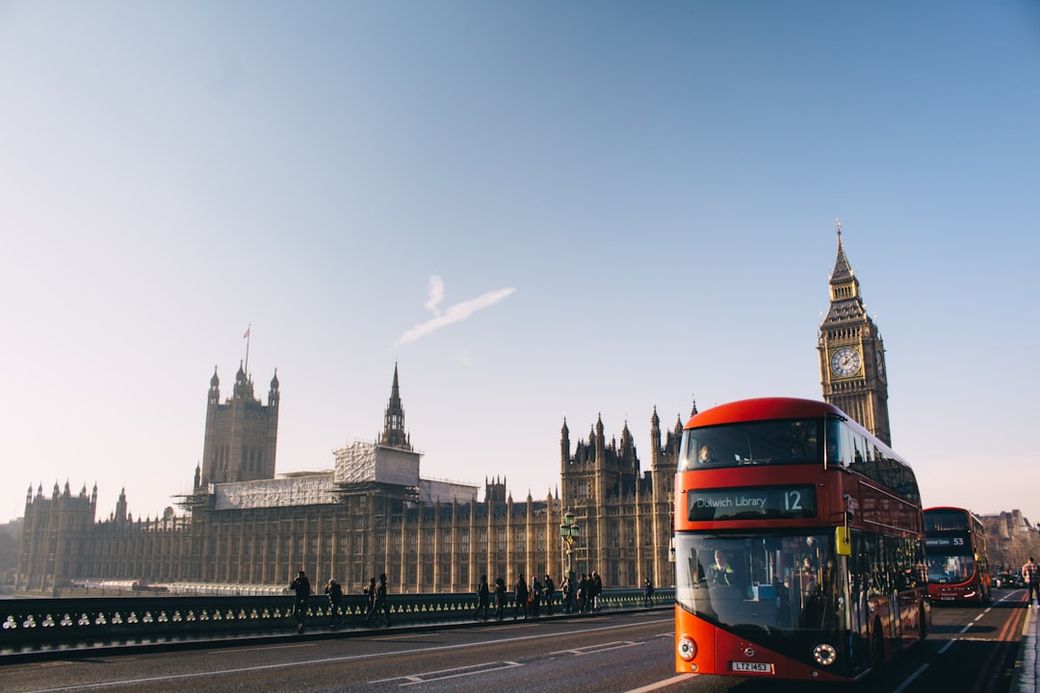 The image depicts a red double-decker bus on a road in front of the iconic Big Ben and the Houses of Parliament in London. The scene captures a clear blue sky, highlighting the architectural beauty of the historical buildings. — Credit: Photo by Aron Van de Pol on Unsplash