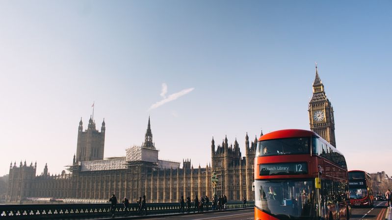 The image depicts a red double-decker bus on a road in front of the iconic Big Ben and the Houses of Parliament in London. The scene captures a clear blue sky, highlighting the architectural beauty of the historical buildings.