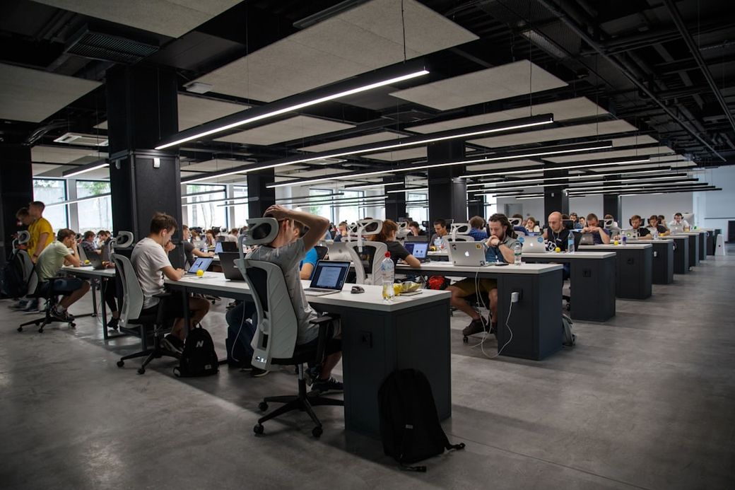 A modern open office space filled with rows of individuals working on computers. The environment showcases collaborative workstations with noticeable engagement among the members. — Credit: Photo by Alex Kotliarskyi on Unsplash